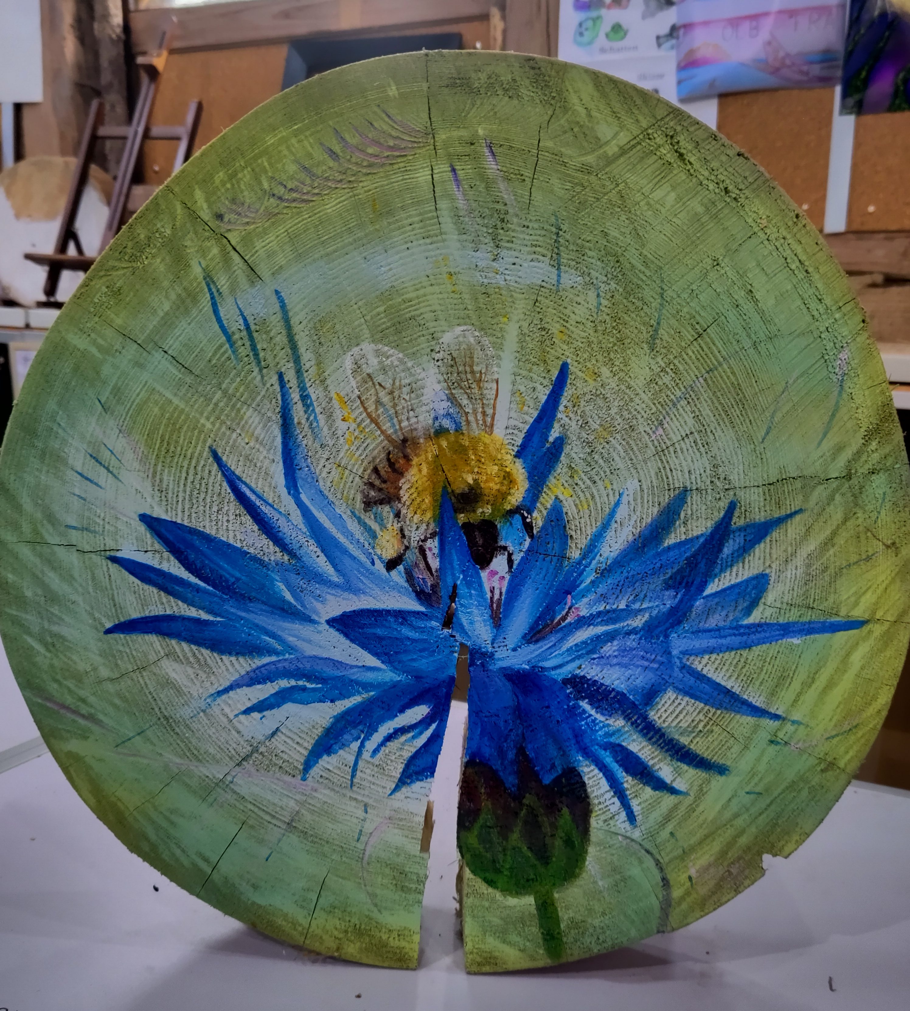 Bemalte Baumscheibe mit blauer Kornblume und Hummel auf grünem Hintergrund